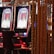 A pair of casino slot machines, each with detailed screens displaying various game outcomes and jackpot amounts. The chairs in front of the slot machines are dark and cushioned for comfort. The background features a hanging curtain made of golden ropes and other casino decorations are visible.