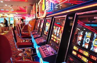 A brightly lit casino interior featuring a row of colorful slot machines. The neon lights create a vibrant atmosphere, with patterned carpeting adding to the decor. Stools are aligned neatly in front of each machine.