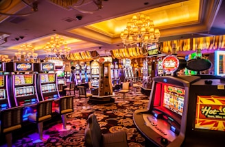 A vibrant casino interior filled with rows of colorful slot machines, each illuminated with bright lights and displays. The ceiling is adorned with elegant chandeliers and golden drapes, creating a luxurious atmosphere. The patterned carpet adds to the elaborate design of the space.