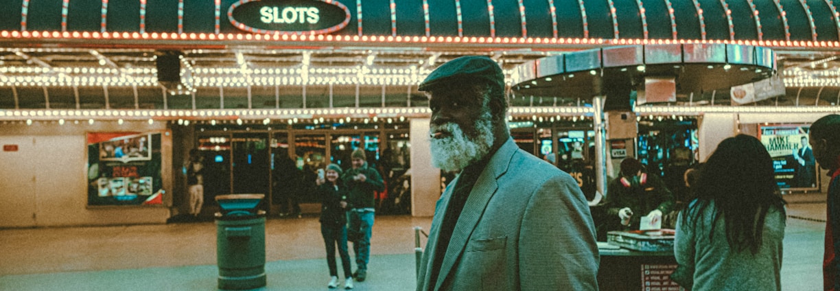 An older man with a gray beard and wearing a suit and hat stands in front of a brightly lit casino. The background features a large illuminated sign reading 'CASINO' and 'SLOTS' among other words, with numerous lights outlining the entrance. Several other people are visible in the background, engaged in various activities, some walking, some standing, and one looking at something on display.