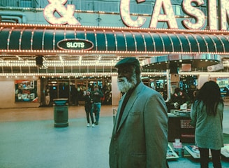 An older man with a gray beard and wearing a suit and hat stands in front of a brightly lit casino. The background features a large illuminated sign reading 'CASINO' and 'SLOTS' among other words, with numerous lights outlining the entrance. Several other people are visible in the background, engaged in various activities, some walking, some standing, and one looking at something on display.
